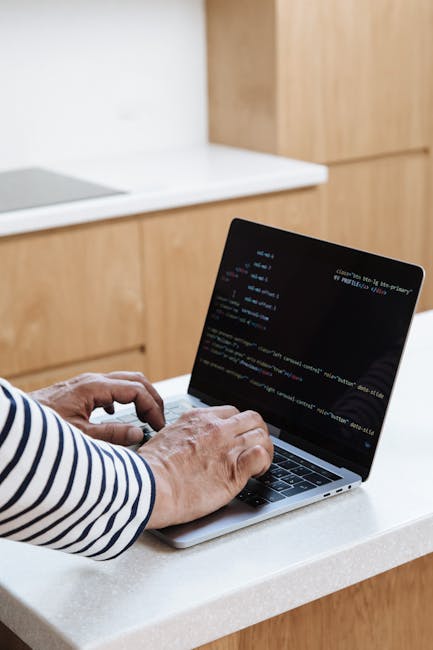 The image features a person wearing a long-sleeved, white and navy striped shirt operating a silver laptop placed on a white kitchen countertop. The laptop's screen displays lines of colored code on a